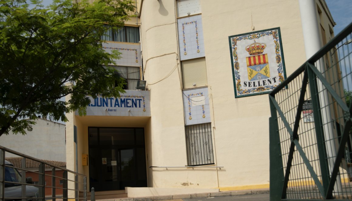 Fachada casi completa del ayuntamiento y entorno urbano con coches aparcados. En la parte derecha, un cuadro de cerámica de gran tamaño muestra el escudo del pueblo.