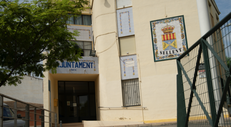 Fachada casi completa del ayuntamiento y entorno urbano con coches aparcados. En la parte derecha, un cuadro de cerámica de gran tamaño muestra el escudo del pueblo.